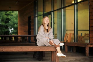 A woman with light brown hair is wrapped in a sweater and sitting cross-legged on a wooden bench.