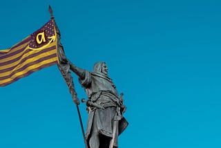“Statue of a medieval knight raising a flag with modern corporate symbols: an ‘a’ with an arrow, set against a blue sky, symbolizing technocapitalism.”