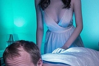 A 21 year old Chinese woman, wearing a thin blue nightgown, massages a naked 35 year old white man, lying on his stomach on a massage table, in a dimly lit massage parlor.