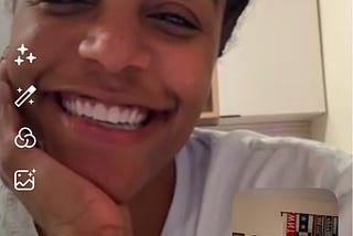 A screenshot of a video call featuring two smiling individuals. The main screen shows a woman with a radiant smile, wearing a white shirt, resting her chin on her hand. The smaller screen in the bottom-right corner displays another smiling person, wearing a plaid shirt, sitting in a room with colorful decorations and posters in the background.