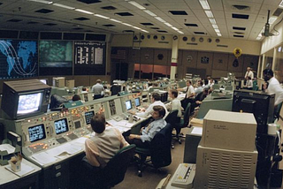 Wide angle view of the flight control room (FCR) of the Mission Control Center (MCC). Some of the STS 41-G crew can be seen on a large screen at the front of the MCC along with a map tracking the progress of the orbiter. It’s pretty intense!
