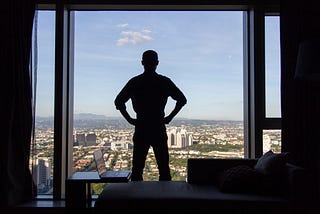 The silhouette of a man with his back turned standing at his full height with his hands on his hips, in front of a floor-to-ceiling window with a city view, with a chaise lounge sofa and an open laptop on a side table behind him.