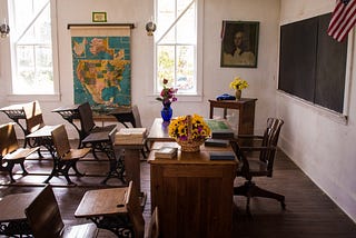 An empty classroom with wooden desks and colorful flowers on the teacher’s desk.
