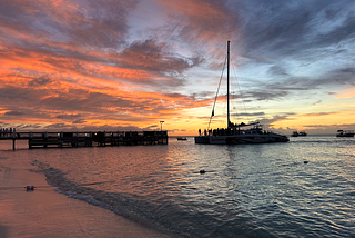 Large catamaran sailboat approaching pier on the beach at sunset.