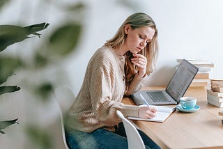 Woman writing at a desk with a laptop, notebook, and coffee.