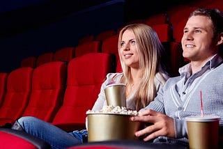 Descrição: foto de um homem e uma mulher que estão sozinhos em uma sala de cinema. A mulher aparenta ser jovem e está usando uma blusa bege clara, ela é branca com cabelo loiro e liso. O homem também aparenta ser jovem, está vestindo uma camisa em um tom de cinza claro e um suéter com um tom de cinza mais escuro por cima, porém com listas brancas ao redor da gola. Ele é branco e seu cabelo é castanho claro. Estão sorrindo e segurando um balde de pipoca com um copo de refrigerante para cada um.