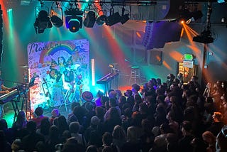 A photo of Madeline the Person performing in Seattle, Washington. Colorful rainbow lights wash over the stage as she sings and strums a guitar in front of a collaged backdrop of her and her fans art.