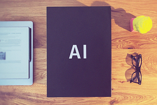 A black box labeled AI on a desk with glasses, a small statue and an e-notebook.