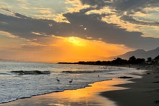 Photo taken by author of a sunset at the beach in Carpinteria, CA. There are clouds across the sky, sun hanging near the horizon in a brilliant orange, reflecting off the sand, and birds in the frothy white surf at the shore, and mountains in the distance. People walk their dogs and play as the sun still casts its light.