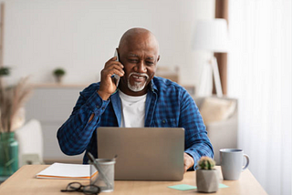 Older man talking on phone while on laptop.