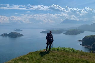 A hiker stands silhouetted against a beautiful scottish coastline shrouded  in brilliant summer haze