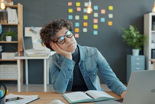 A writer at her desk falling asleep out of boredom. She’s wearing a blue jacket and her laptop is open on the desk.