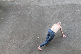 A person in blue-grey jeans walks around on all fours like a dog. They are shirtless, and the ground beneath them is concrete. They have their back to us and we cannot see their face.