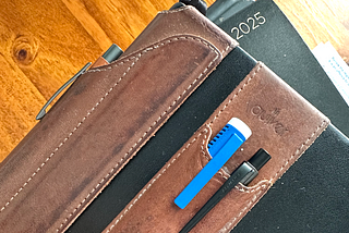 Black notebook, closed, with two tan leather pen holders strapped to the outside. there’s a bright blue eraser pen with a white cap and a mechanical pencil in the double pen holder on the front of the book, and a tan coloured clicky ballpoint pen in the holder that cradles the spine.