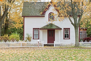 A white house with a red door, gray-green roof, with a white picket fence and a tree outside in fall with the yellow leaves on the ground