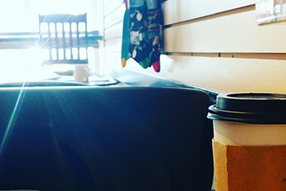 A photo of a lap top next to a cup of take out coffee, in a coffee shop, with sunlight streaming in through the window