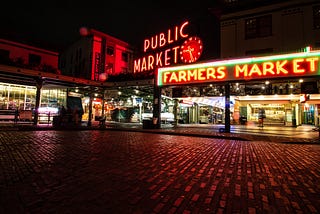 The neon signs of Seattle’s Pike Place Market illuminated at night. They read, “Public Market” and “Farmers Market”.
