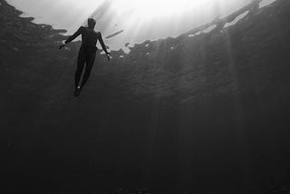 Person in water, seen from underneath. Their head is at the surface and their feet are well above the ocean floor. Black and white, with rays of light shining from above.