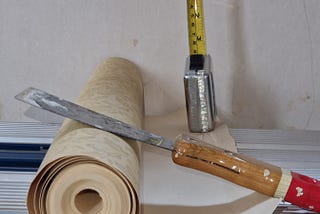 A coloured image showing a roll of wallpaper, a tape measure and a wallpaper scraper sitting on a metal bench against a bare wall background.