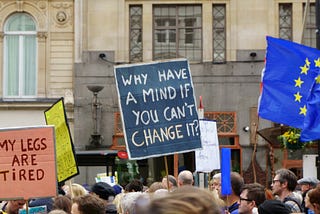 Photo of a protest. There people holding up signs which read (from left to right): “My legs are tired” and “Why have a mind if you can’t change it?”