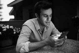 A man sitting at a table looks at a mobile phone that he’s holding in his hands.