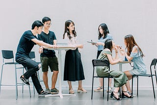 Two men and four women standing and sitting, working and talking together.