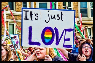 A group of people carrying signs and flags. In the foreground, the camera is focused on a sign that says, “It’s just LOVE.” The centre of the O is filled in with rainbow colours.