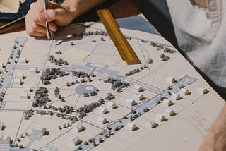 A man holding a pen and working on a scale model of an architectural plan, with tiny trees and sketches on the paper.
