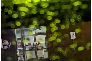 Image shows neon green “Planet Fitness” sign fractallated by raindrops collected on automobile glass.