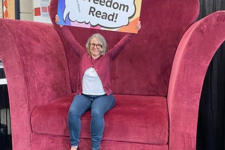 Librarian sitting on an oversized chair holding a sign saying, “Let Freedom Read!”