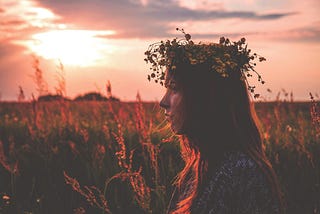 Warm orange and pink silhouette of a woman wearing a floral wreath, standing in a cornfield at sunset.