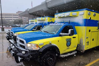 Two ambulances are parked outside a station during a snow storm.
