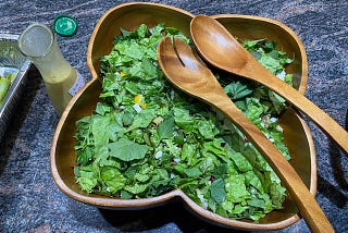 Mixed green salad in clover-leaf wooden bowl with wooden serving utensils and lemon dressing in a pouring container