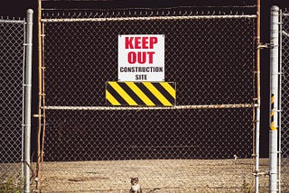 A cat sits behind a fence, peering through a gate with a sign that warns “keep out”