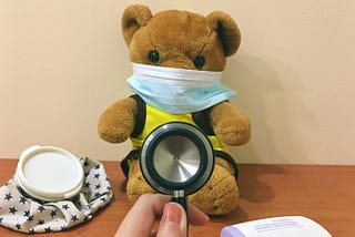 Photograph of brown teddy bear wearing a blue surgical mask while a doctor’s hands hold a stethoscope in front of it.