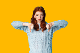 Red haired young woman in a light blue jumper with her arms raised in front of her, with her two thumbs pointing down. She looks a bit negative. Bright yellow background