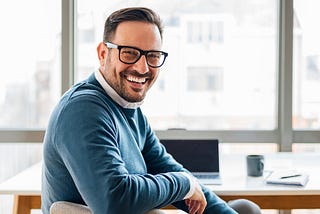 A writer smiling while sitting at a wooden desk. In the background, a laptop, coffee mug, and notebook are visible.