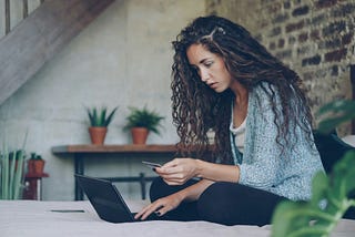 A woman sitting on a bed with an open laptop, holding a credit card.