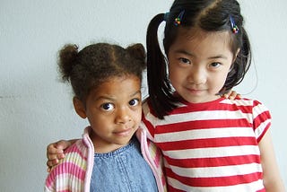 A Black child and Asian child facing the camera in a partial embrace against an off-white background. The Black child is a little girl with pigtails, wearing a blue shirt with a pink-and-white stripped cardigan. The taller Asian child is also wearing pigtails and is wearing a red-and-white stripped shirt.