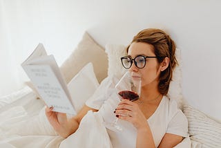 Woman in White Shirt Holding a Glass of Wine reading a book in bed.