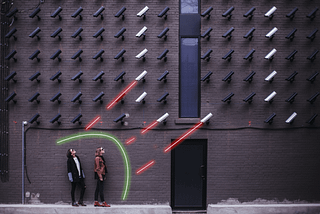 Two women, shielded by a great laser and in front of security cameras which shoot red lasers at them.