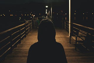 A person wearing a hoodie in a shadow, standing on a boardwalk at night.