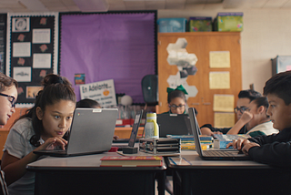 A group of elementary students in a classroom looking at laptops in their desks.