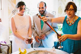 A couple holds items for the ritual end of the Sabbath as Rabbi Nikki, a white woman in glasses and bright clothing, extinguishes the braided candle in wine.