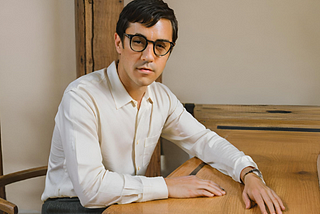 A neatly-dressed man with black hair and glasses sitting at a desk.