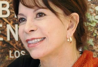 Isabel Allende is smiling and looking into the distance. She is wearing a black top, partially covered by an orange scarf.