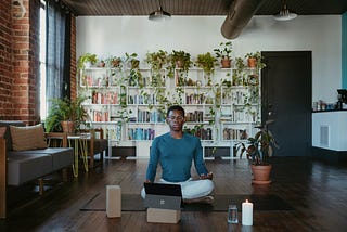 Man meditating in a room filled with books and plants and a burning candle.