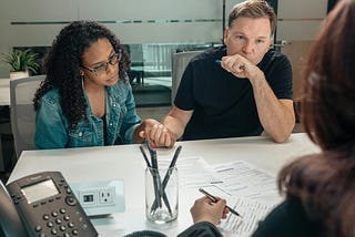 An adult couple sits holding hands across the desk from a doctor. On the desk between them are papers, pens and a phone.