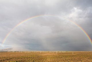 A large rainbow over a cacti filled desert scene with clouds and mountains in the background