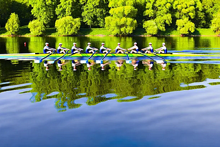 AI generated image of a crew of rowers. Note that some folks are backwards from what they should be. They are rowing on a still lake or river lined with green trees. The boat is bright green with the rowers in blue bibs and white undershirts. The oars are blue. The reflection of the trees is clear in the still water.